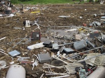 An outdoor scene filled with scattered debris and discarded electronic waste. The ground is covered with broken metal pieces, wires, and various components, creating a cluttered and chaotic atmosphere. In the background, an area with green vegetation is visible.