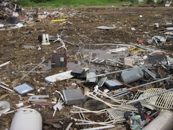 An outdoor scene filled with scattered debris and discarded electronic waste. The ground is covered with broken metal pieces, wires, and various components, creating a cluttered and chaotic atmosphere. In the background, an area with green vegetation is visible.