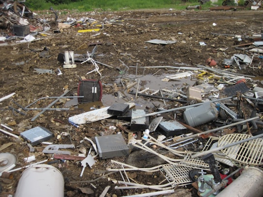 An outdoor scene filled with scattered debris and discarded electronic waste. The ground is covered with broken metal pieces, wires, and various components, creating a cluttered and chaotic atmosphere. In the background, an area with green vegetation is visible.