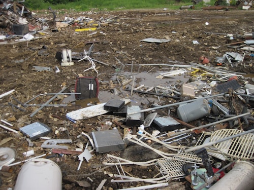 An outdoor scene filled with scattered debris and discarded electronic waste. The ground is covered with broken metal pieces, wires, and various components, creating a cluttered and chaotic atmosphere. In the background, an area with green vegetation is visible.