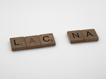 Wooden letter tiles are arranged on a white surface, spelling out two sets of letters: L, A, C, and N, A. Each tile has a single letter and a small number indicating its value.