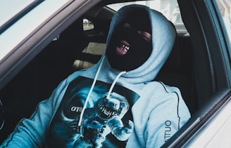 A person is sitting in a car wearing a black ski mask and a gray hoodie featuring a graphic of an astronaut. The individual is showing grills on their teeth and appears relaxed or amused.