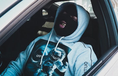 A person is sitting in a car wearing a black ski mask and a gray hoodie featuring a graphic of an astronaut. The individual is showing grills on their teeth and appears relaxed or amused.