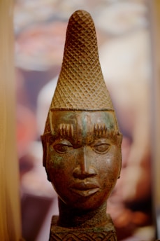 An intricately carved sculpture head with a tall, textured headdress, smooth facial features, and pronounced eyebrows. The sculpture exhibits a blend of earthy tones with a subtle patina that suggests age and craftsmanship.