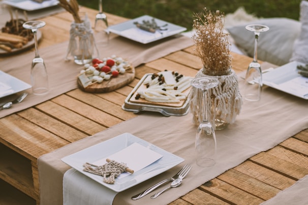 A rustic wooden table is set outdoors with a beige table runner. The table features white square plates, neatly folded napkins with decorative twine, and upside-down wine glasses. There are appetizers on wooden boards, including slices of cheese and small tomatoes. A decorative jar with dried flowers adds a natural touch.