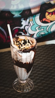 A tall, clear glass filled with a chocolate milkshake topped with whipped cream, chocolate syrup, nuts, and chocolate chips. A white straw is placed in the milkshake. The glass is placed on a dark woven surface. In the background, there is a colorful, patterned design.