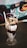 A tall, clear glass filled with a chocolate milkshake topped with whipped cream, chocolate syrup, nuts, and chocolate chips. A white straw is placed in the milkshake. The glass is placed on a dark woven surface. In the background, there is a colorful, patterned design.