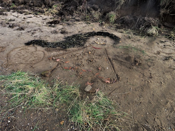 A drawing on the ground made with natural materials such as grass, twigs, and stones. It appears to be a figure with a smiley face and hair made of dark vegetation. There's a heart-shaped drawing nearby with writing etched into the earth. The surrounding area is muddy with patches of grass and some fallen leaves.