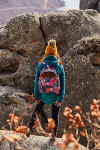 A person dressed in outdoor clothing, including a teal jacket and a mustard yellow knitted hat with a pom-pom, stands on rocky terrain. A colorful floral backpack is visible, and the person appears to be looking at a large rock formation. Dried plants with orange leaves are in the foreground, and a mountainous landscape is in the background.