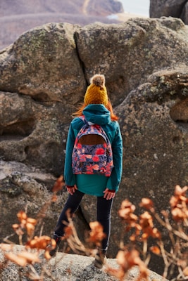 A person dressed in outdoor clothing, including a teal jacket and a mustard yellow knitted hat with a pom-pom, stands on rocky terrain. A colorful floral backpack is visible, and the person appears to be looking at a large rock formation. Dried plants with orange leaves are in the foreground, and a mountainous landscape is in the background.