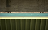 A close-up view of a bridge structure featuring a concrete support and a metal beam with a teal colored surface in between. The photo highlights the geometric patterns and textures of the materials used in the construction.