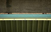 A close-up view of a bridge structure featuring a concrete support and a metal beam with a teal colored surface in between. The photo highlights the geometric patterns and textures of the materials used in the construction.