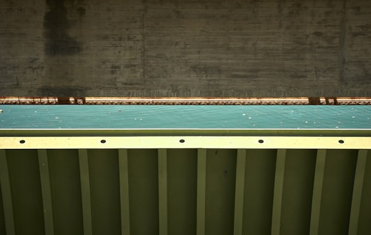 A close-up view of a bridge structure featuring a concrete support and a metal beam with a teal colored surface in between. The photo highlights the geometric patterns and textures of the materials used in the construction.