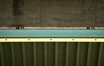 A close-up view of a bridge structure featuring a concrete support and a metal beam with a teal colored surface in between. The photo highlights the geometric patterns and textures of the materials used in the construction.