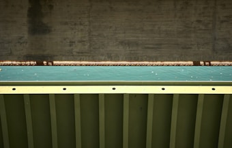 A close-up view of a bridge structure featuring a concrete support and a metal beam with a teal colored surface in between. The photo highlights the geometric patterns and textures of the materials used in the construction.