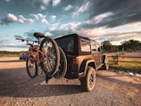 A rugged off-road vehicle is parked in a sunlit area with two bicycles mounted on a rear bike rack. The sky is partly cloudy with a mix of white, fluffy clouds against a backdrop of blue. Trees and an open field are visible in the distance.
