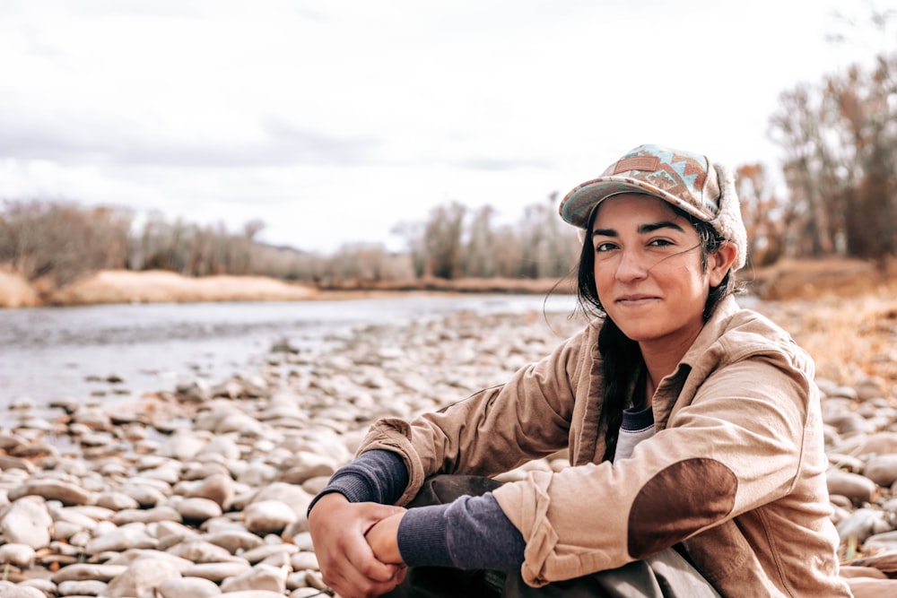 woman in brown jacket and blue denim jeans sitting on brown rock near body of water