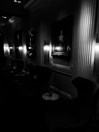 A dimly lit room features a series of framed portraits on the walls. Elegant armchairs and small round tables are arranged along the side. Art deco-style wall lights provide a soft, ambient glow.