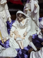 A ceramic nativity scene featuring a figure of a woman with brown hair wearing a white robe, sitting with an infant in her lap. Surrounding them are other figures, including a standing person holding a gift. The background includes a deep purple fabric draped underneath the figures.