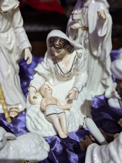 A ceramic nativity scene featuring a figure of a woman with brown hair wearing a white robe, sitting with an infant in her lap. Surrounding them are other figures, including a standing person holding a gift. The background includes a deep purple fabric draped underneath the figures.