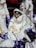 A ceramic nativity scene featuring a figure of a woman with brown hair wearing a white robe, sitting with an infant in her lap. Surrounding them are other figures, including a standing person holding a gift. The background includes a deep purple fabric draped underneath the figures.