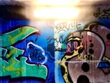 A vibrant graffiti mural on a concrete wall illuminated by an overhead fluorescent light. The artwork features abstract shapes and bold colors with a combination of green, blue, and black swirls. Some letters and doodles blend into the background, and the overall style is expressive with spray-painted layers.