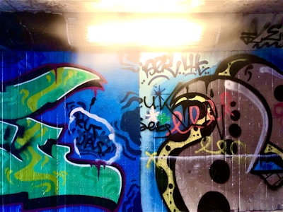 A vibrant graffiti mural on a concrete wall illuminated by an overhead fluorescent light. The artwork features abstract shapes and bold colors with a combination of green, blue, and black swirls. Some letters and doodles blend into the background, and the overall style is expressive with spray-painted layers.