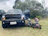 A large black Ford pickup truck with its front doors open is parked on grass. In front of the truck are two small quad bikes, one blue and one pink with helmets placed on their seats. The background consists of trees and cloudy sky.