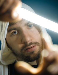 A person with a focused expression holds a glowing light source close to their face. They are wearing a hooded white garment, and their blue eyes are prominently visible. The background is blurred, emphasizing the person's face and the bright light.