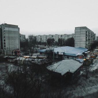 A residential urban area features several large, concrete apartment blocks surrounding a central structure. The scene is overcast with a gloomy atmosphere, and bare trees are visible among the buildings. The setting appears somewhat neglected, and the ground is wet, suggesting recent rain.