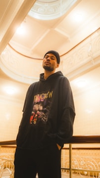 A person stands indoors wearing a black hoodie with 'DJ Screw' printed on it. The setting appears elegant, with ornate architectural details and warm lighting creating a soft, inviting atmosphere.