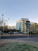 An urban street scene featuring a multi-story residential building with a blue and beige facade. Several parked cars are in a lot adjacent to the building. A street sign labeled 'Mühlenweg' is visible in the foreground, along with some trees and a lamp post. The sky is clear and blue.