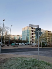 An urban street scene featuring a multi-story residential building with a blue and beige facade. Several parked cars are in a lot adjacent to the building. A street sign labeled 'Mühlenweg' is visible in the foreground, along with some trees and a lamp post. The sky is clear and blue.