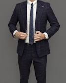 A person wearing a dark blue checkered suit with a white shirt and a matching blue tie. The hands are adjusting the button of the suit jacket. The background is plain gray.