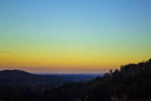 A serene landscape depicting a silhouetted forest with gentle hills in the foreground, under a gradient sky transitioning from blue to yellow, suggesting a sunset or sunrise.