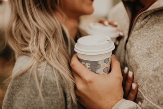 Two people are close together, each holding a to-go coffee cup with white lids. They are wearing cozy, neutral-colored clothing, and one person has long, blond hair. The scene suggests warmth and intimacy.
