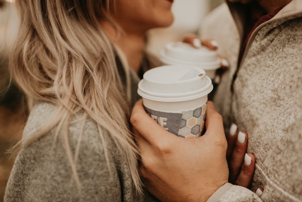 Two people are close together, each holding a to-go coffee cup with white lids. They are wearing cozy, neutral-colored clothing, and one person has long, blond hair. The scene suggests warmth and intimacy.