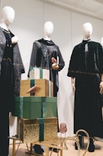 An arrangement of mannequins dressed in elegant black clothing with ornate accessories, placed behind a stack of decorative gift boxes. The boxes are adorned in green and gold wrapping, topped with small metallic bird ornaments. The scene is set on a light wooden floor, with a calm and sophisticated ambiance.