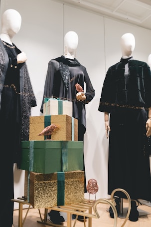 An arrangement of mannequins dressed in elegant black clothing with ornate accessories, placed behind a stack of decorative gift boxes. The boxes are adorned in green and gold wrapping, topped with small metallic bird ornaments. The scene is set on a light wooden floor, with a calm and sophisticated ambiance.