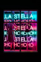 A brightly lit store window display featuring a mannequin dressed in a stylish outfit. The background is adorned with vivid neon lights spelling out 'LA STELLA' and 'HO HO HO' in alternating bright hues of blue, pink, and red. The overall atmosphere is vibrant and colorful, suggesting a festive or celebratory theme.