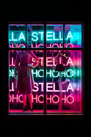 A brightly lit store window display featuring a mannequin dressed in a stylish outfit. The background is adorned with vivid neon lights spelling out 'LA STELLA' and 'HO HO HO' in alternating bright hues of blue, pink, and red. The overall atmosphere is vibrant and colorful, suggesting a festive or celebratory theme.