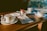 A wooden table is set with white ceramic cups and saucers, along with plates and napkins. A small container in the center holds various packets, possibly of sugar or creamer. The sunlight casts warm tones across the table, and the background suggests a view of a forest or greenery outside.