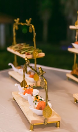 A set of elegantly plated appetizers displayed on wooden trays with decorative gold-colored stands. The dishes are presented on small white boat-shaped dishes, containing portions of salmon garnished with herbs. The setting appears to be outdoors, possibly at a catered event or upscale gathering.