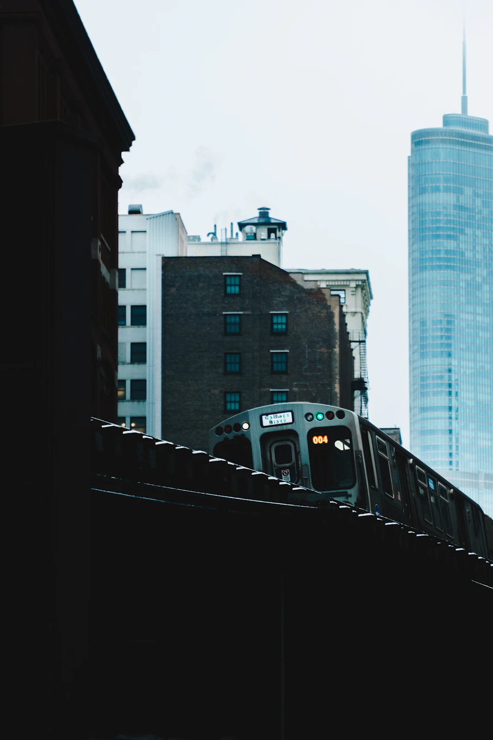 Train crosses city street under overcast sky with contrasting blue-white locomotive and buildings
