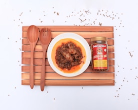A wooden tray with two wooden utensils, a jar of spicy sauce, and a plate containing a spicy dish. The dish appears to have pieces of seafood in a rich, oily sauce. The jar next to the plate has a label indicating the contents are a type of chili sauce.