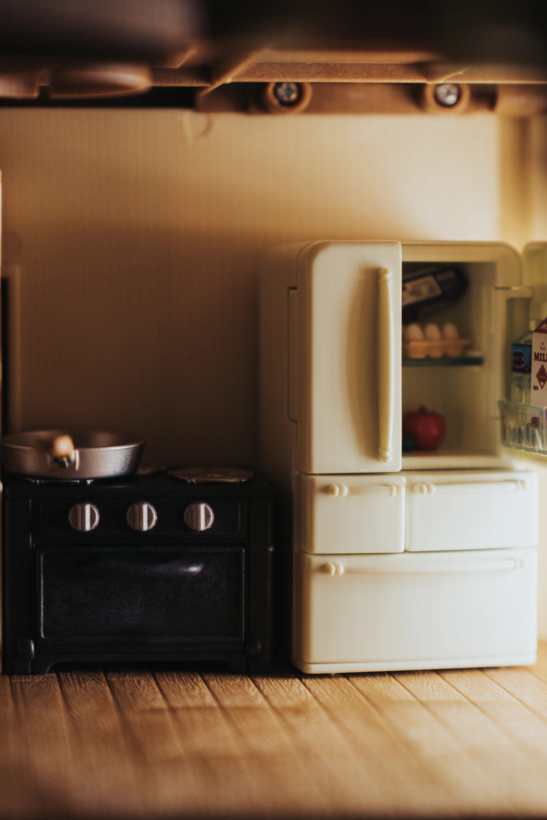 A miniature kitchen with a white refrigerator standing open beside a black stove on a wooden floor