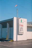 A large billboard on the side of a building displays a Coca-Cola advertisement with the phrase 'Coke adds life to... Nashville.' The building is constructed with white bricks and has an industrial appearance. A windsock in orange is mounted on a pole above the billboard, and the sky is clear and blue.