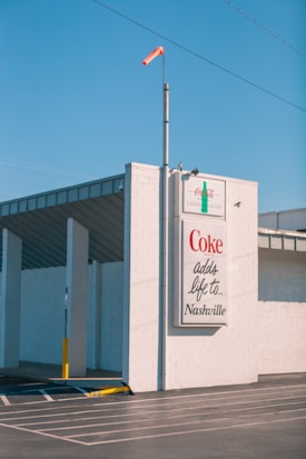 A large billboard on the side of a building displays a Coca-Cola advertisement with the phrase 'Coke adds life to... Nashville.' The building is constructed with white bricks and has an industrial appearance. A windsock in orange is mounted on a pole above the billboard, and the sky is clear and blue.