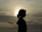 A silhouette of a person with curly hair is visible against a backdrop of a dramatic sky. The sun is partially obscured by clouds, casting a gentle glow around the figure. The scene is framed with what appears to be a fence in the foreground, adding depth to the image.