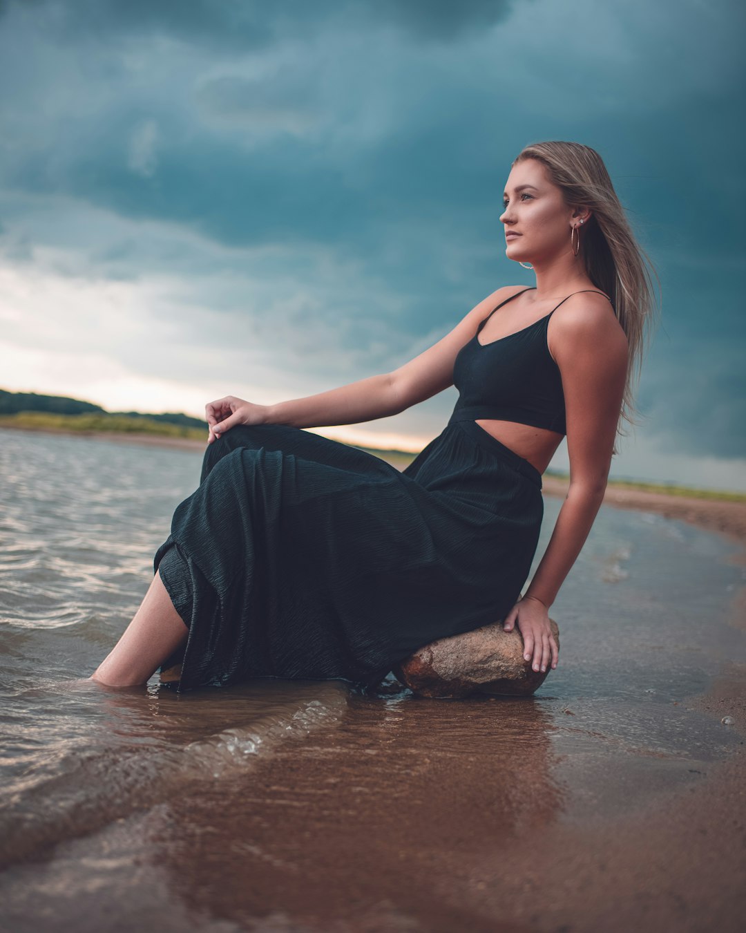 woman in black spaghetti strap dress sitting on brown rock on water during daytime